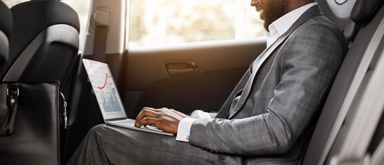 Smiling black guy businessman working on laptop, reading marketing report while going by car to business meeting. African man in suit sitting on back car seat, typing on laptop keyboard, side view