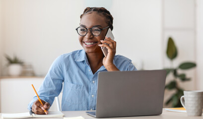 Smiling Black Female Office Manager Talking On Cellphone And Taking Notes At Workplace, Wearing...