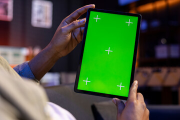 Young remote worker uses green screen on a tablet at his house, feeling carefree in the living room and solving his freelancing tasks. Male person being casual in his apartment, teleworking.