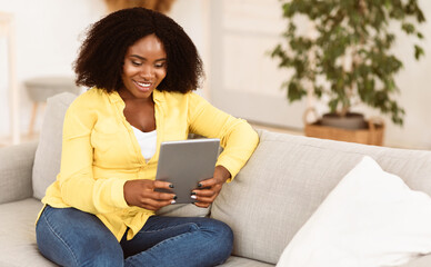 Smiling black curly girl holding tablet, sitting on couch in light living room in her modern...