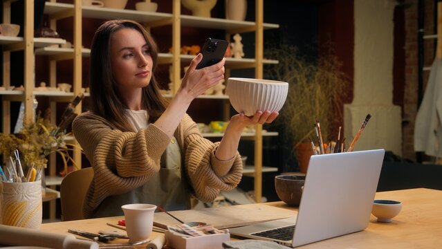 Caucasian woman girl female potter sculptor artist blogger using mobile phone handmade bowl photo in workshop crafting hobby small business owner sale ceramics online shop pottery selling by laptop - Powered by Adobe