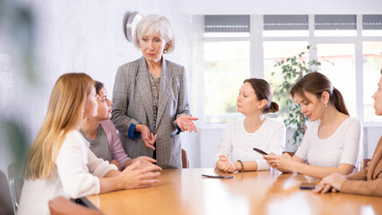 during morning five minutes, colleagues sit at table and animatedly discuss set of tasks for day. Senior woman team leader stands and leads staff meeting.Group work, teamwork
