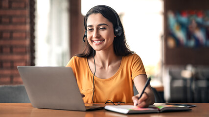 Happy young arab woman having educational course online, using laptop and headset, cafe interior, free space. Smiling female student studying online, sitting in front of laptop at cafeteria