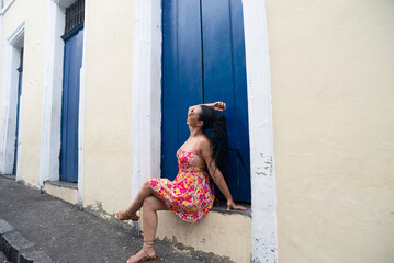 Beautiful young Caucasian woman with braids and colorful dress sitting in front of a blue door of...