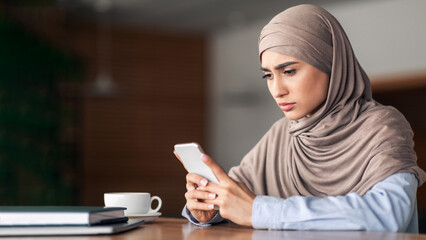 Sad muslim girl holding mobile phone, cafe interior, empty space