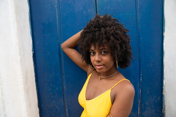 Fototapeta premium Close-up portrait of a young woman with afro hair, wearing a yellow dress, sitting by a blue door of an old house, looking at the camera. Confident person.