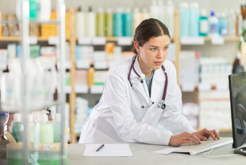 Confident skilled young female drugstore staff member in white coat inputting purchase orders into...