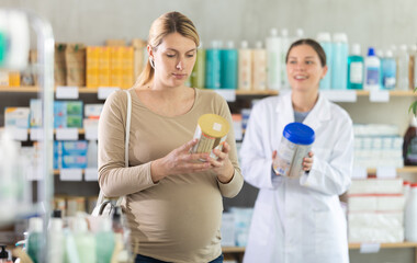 Pregnant woman choosing baby formula with help of young female pharmacist in pharmacy