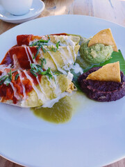 Dobladas de queso bañadas con salsa roja y crema blanca, acompañadas de frijoles negros, guacamole y nachos. Toma vertical.