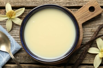 Tasty condensed milk, spoon, vanilla pods and flowers on wooden table, flat lay