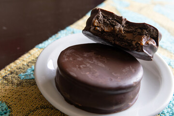 A dark chocolate alfajor inside a small plate with another half-finished alfajor showing the dulce de leche filling