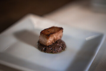 Seared pork belly on dark sauce, presented on a square white plate. Minimalistic fine dining style with blurred background in an elegant setting.