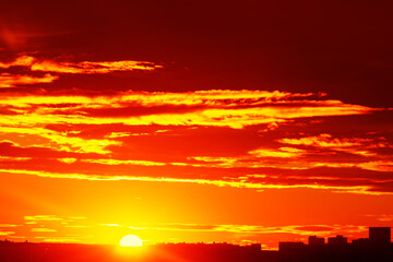 Radiant sunset spilling across the sky, creating a dramatic and warm atmosphere. Silhouettes of buildings at bottom of frame contrast against the vivid colors, emphasizing beauty of glowing horizon