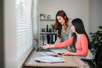 Obraz premium Two women working together at a desk, discussing documents and using a laptop in a bright, modern office environment.