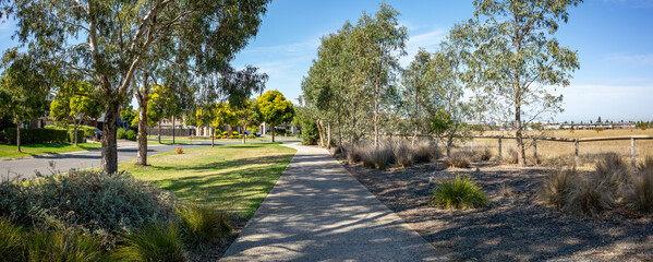 A footpath or sidewalk in a suburban neighborhood with large outdoor space and spacious nature strip. A shade walkway lined with trees and houses in Williams Landing, Melbourne, Australia.