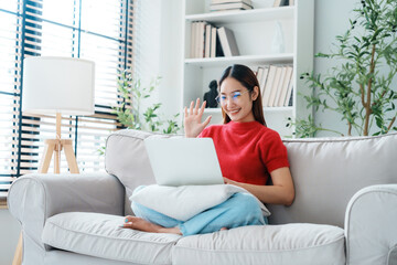 Obraz premium Happy young Asian woman wearing eyeglasses using laptop while seated on couch at home