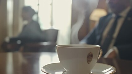 The camera captures a steaming cup of coffee in the foreground with two diplomats engaged in low conversation in the background. Their body language with leanedin postures and intense