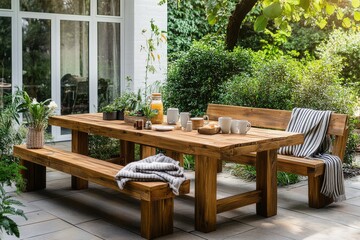 Outdoor Wooden Dining Set with Chairs and Bench on Concrete Patio Surrounded by Greenery. Includes Comfort Items for Gatherings like Coffee Mugs, Blanket, and Pillows.