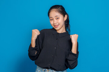 Cheerful young Asian woman smiling brightly with both fists raised in a victorious pose. Her...