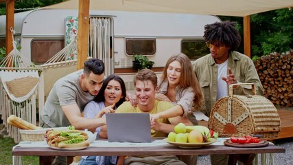 A diverse group of friends relaxes outdoors in a camping setting, sharing laughter while looking at a laptop, have video call. The atmosphere is filled with joy and camaraderie on a warm summer day.