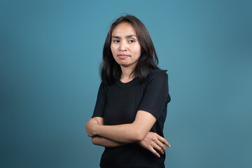 An Indonesian woman in a black shirt crosses her arms while looking away with a displeased or skeptical expression. Posed against a plain blue background, she appears doubtful or unimpressed.