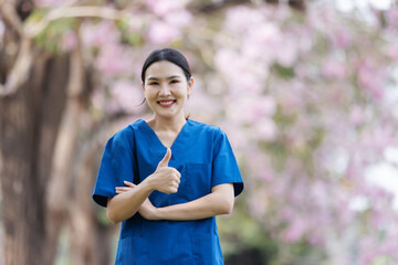 A smiling nurse in blue scrubs gives a thumbs-up gesture while standing in a park filled with blooming trees. The image symbolizes approval, confidence, positivity, and dedication in healthcare.