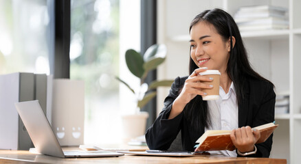Young asian businesswoman drinking coffee, relaxing while working on digital tablet at office desk
