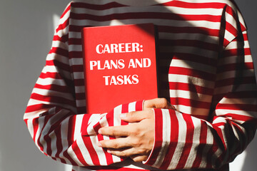 Person woman in red and white striped shirt holding a red notebook with the words "Career: Plans and Tasks" in sunlight, close-up view, concept of professional development