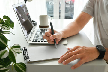 A man is writing in a notebook while sitting at a desk. He is using a pen and the notebook is open to a blank page.