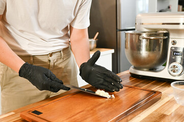 A person is cutting food on a wooden cutting board. The person is wearing black gloves and a white shirt.