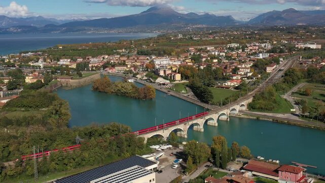 Red high-speed train on the bridge. High-speed train passes an arched railway bridge over a river Mincio in the background Lake Garda Italy.