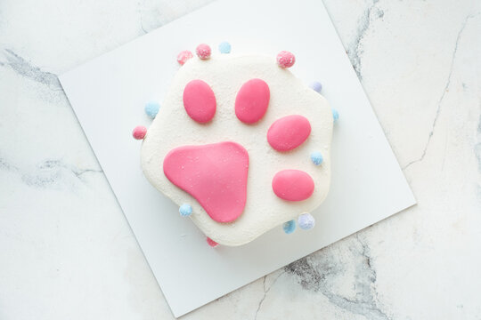 Birthday cake in the shape of trendy squish squishy paw on the white marble background. Flat lay of pink Taba paw shaped cake for a child with white cream cheese frosting