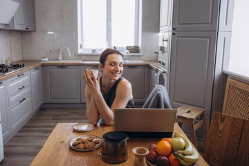A beautiful brunette woman is in the kitchen at the table working on a laptop