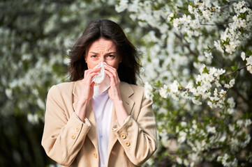 Woman allergic suffering from seasonal allergy at spring in blossoming garden at springtime. Young woman sneezing and blowing nose with nasal handkerchief in front of blooming tree. Allergy concept.