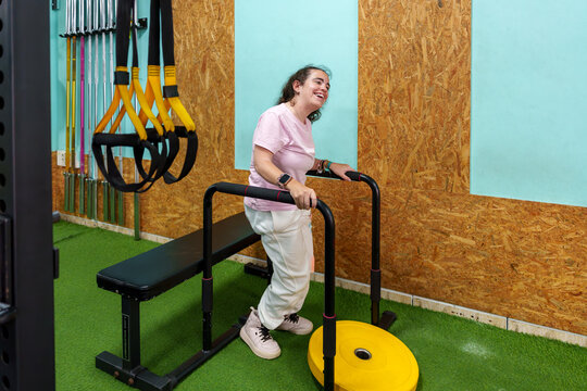 Woman with cerebral palsy using parallel bars, smiling during strength training and physical therapy, demonstrating athletic determination