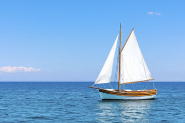 Classic sailboat with white sails glides on tranquil blue ocean under clear sky, sunlight reflecting off its wooden hull and creating peaceful, serene atmosphere
