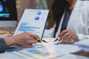 Two businesswomen collaborating on a project, analyzing financial charts and graphs on a table while pointing at data and holding a clipboard with additional information