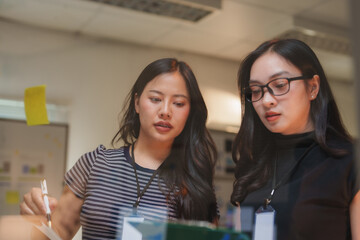 Two young businesswomen are analyzing data and discussing new project ideas, collaborating on a transparent board with sticky notes in a modern office environment