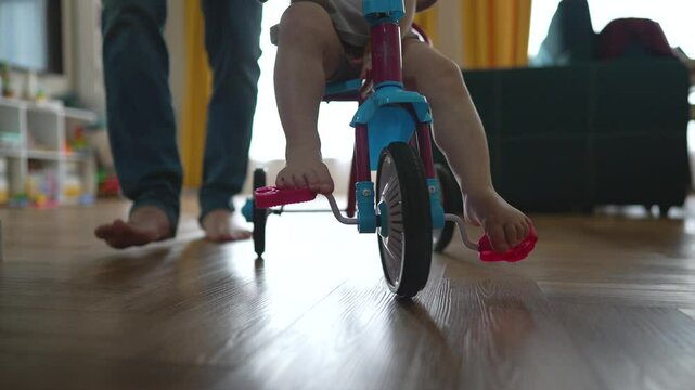 Father guides child on tricycle, playtime indoors. First tricycle ride child pedals with father help. Family fun, learning to ride inside, father support. Dad plays with daughter at home. Happy family