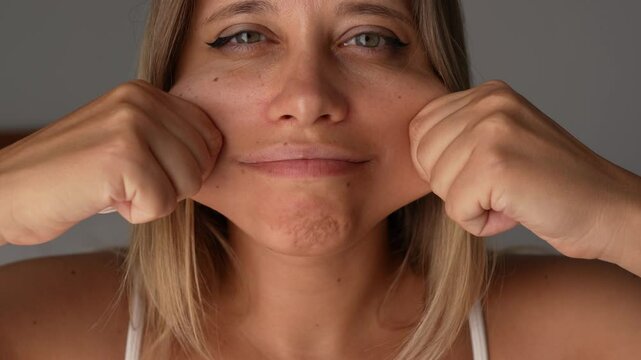 Close-up of a young blonde woman grabbing her chubby cheeks with a smile. A playful and loving moment of self-acceptance, body positivity and joyful connection with her own face