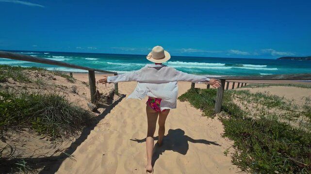 A young woman in a hat and shirt walks along a charming sandy path toward the ocean, surrounded by lush tropical greenery under blue skies.