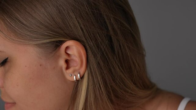 Close-up of young blonde woman showing multiple silver earrings on one ear tucking her hair behind the ear. A modern, stylish detail highlighting personal expression and individuality through jewelry