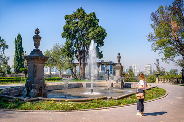 Fototapeta premium A serene scene in Mexico City featuring a historic stone fountain, lush greenery, and a woman looking at the view with modern city buildings in the background