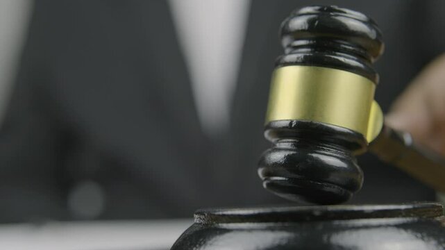 A close-up of a gavel in the foreground, with a person in a black suit signing a document in the background and then striking the gavel, symbolizing the finalization of a legal agreement or decision