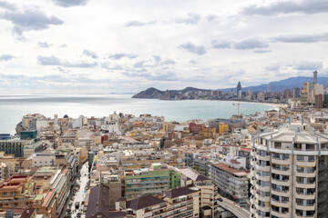 Aerial photo of the Spanish town of Benidorm in Spain showing the old town of the city with the beach known as Platja de Ponent or West Beach Promenade in background as well as mountains and clouds