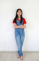 Smart Asian girl child crossed arms with looking camera while standing against white wall in home. Kid in dungarees with Confident gesture.