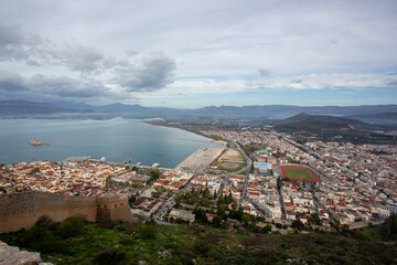 A wide panoramic view of Nafplio city and the Argolic Gulf, seen from the historic Palamidi Fortress in Greece. The coastal city spreads along the shoreline with mountains in the background and a clou