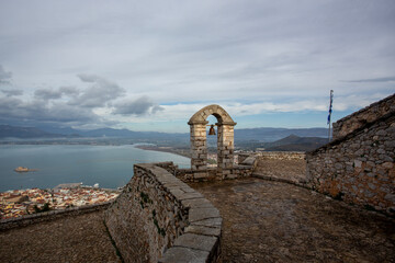 A stone arch with a hanging bell overlooks a panoramic view of the bay and distant mountains under a cloudy sky. Captured from the historic Palamidi Fortress in Nafplio, Greece, this image blends anci