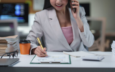 Fototapeta premium Asian businesswoman analyzing financial charts and taking notes while talking on mobile phone, working late at night in office with laptop and paperwork