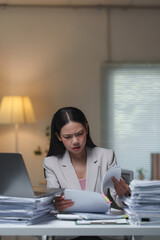 Young businesswoman looking stressed while reviewing documents in a cluttered office at night,...